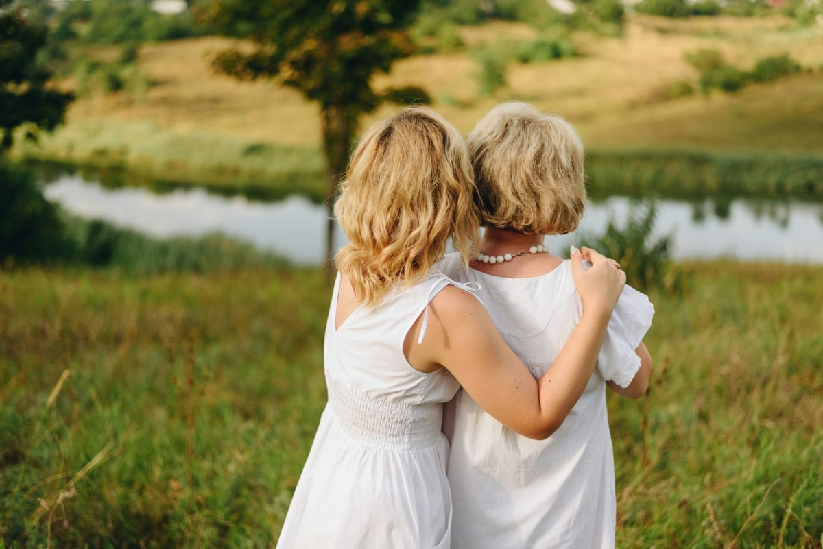 Mãe e filha abraçadas de costas, olhando um lago no campo